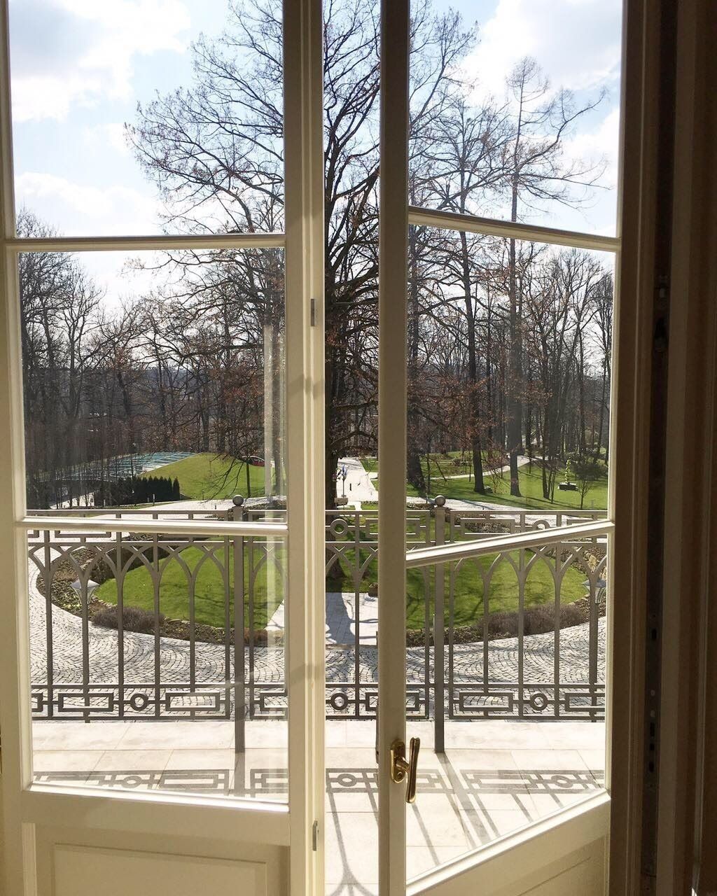 A view from a window with a metal railing, looking out to a park with trees and a grassy area.
