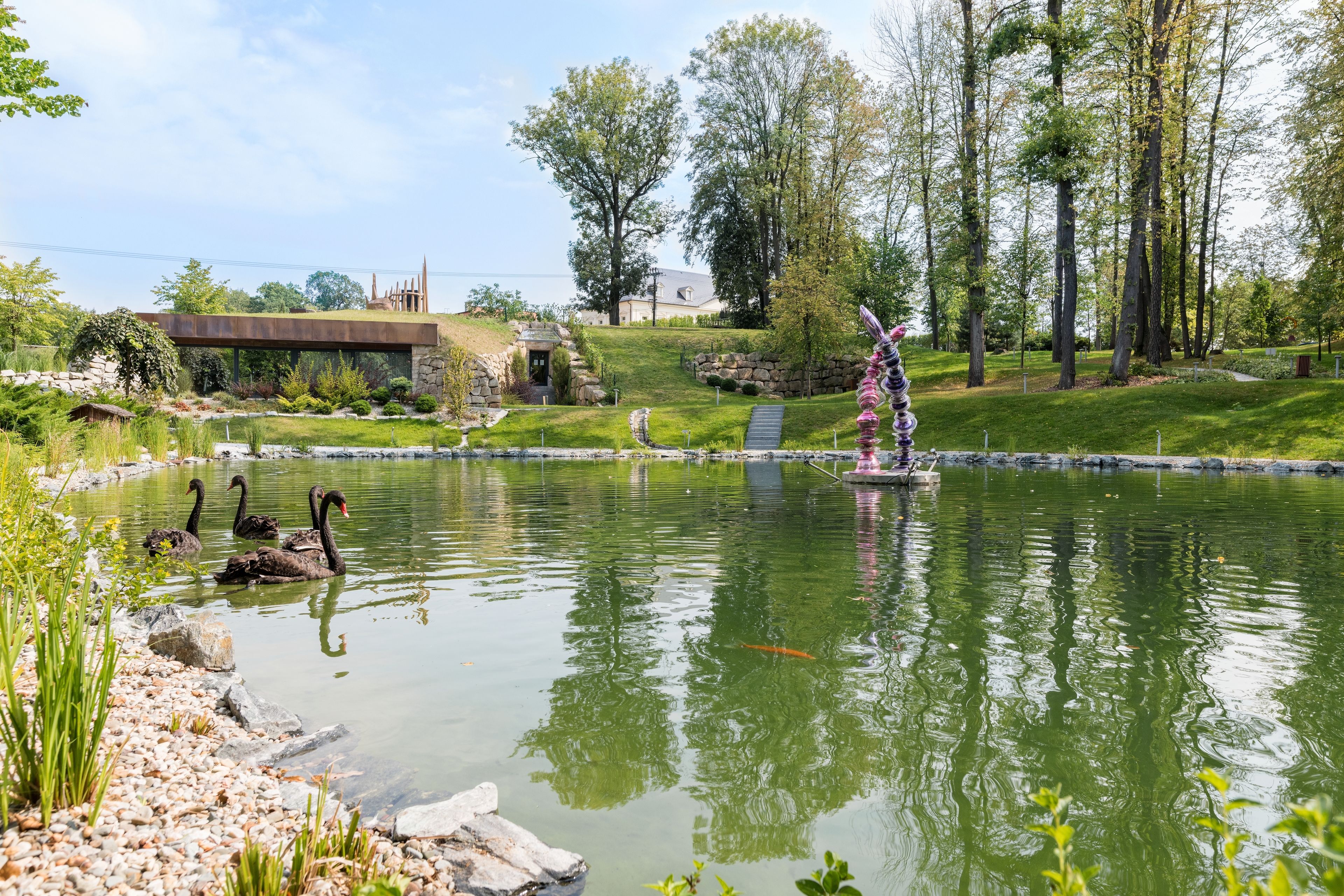 A pond with swans and a statue, surrounded by greenery and a stone bridge.