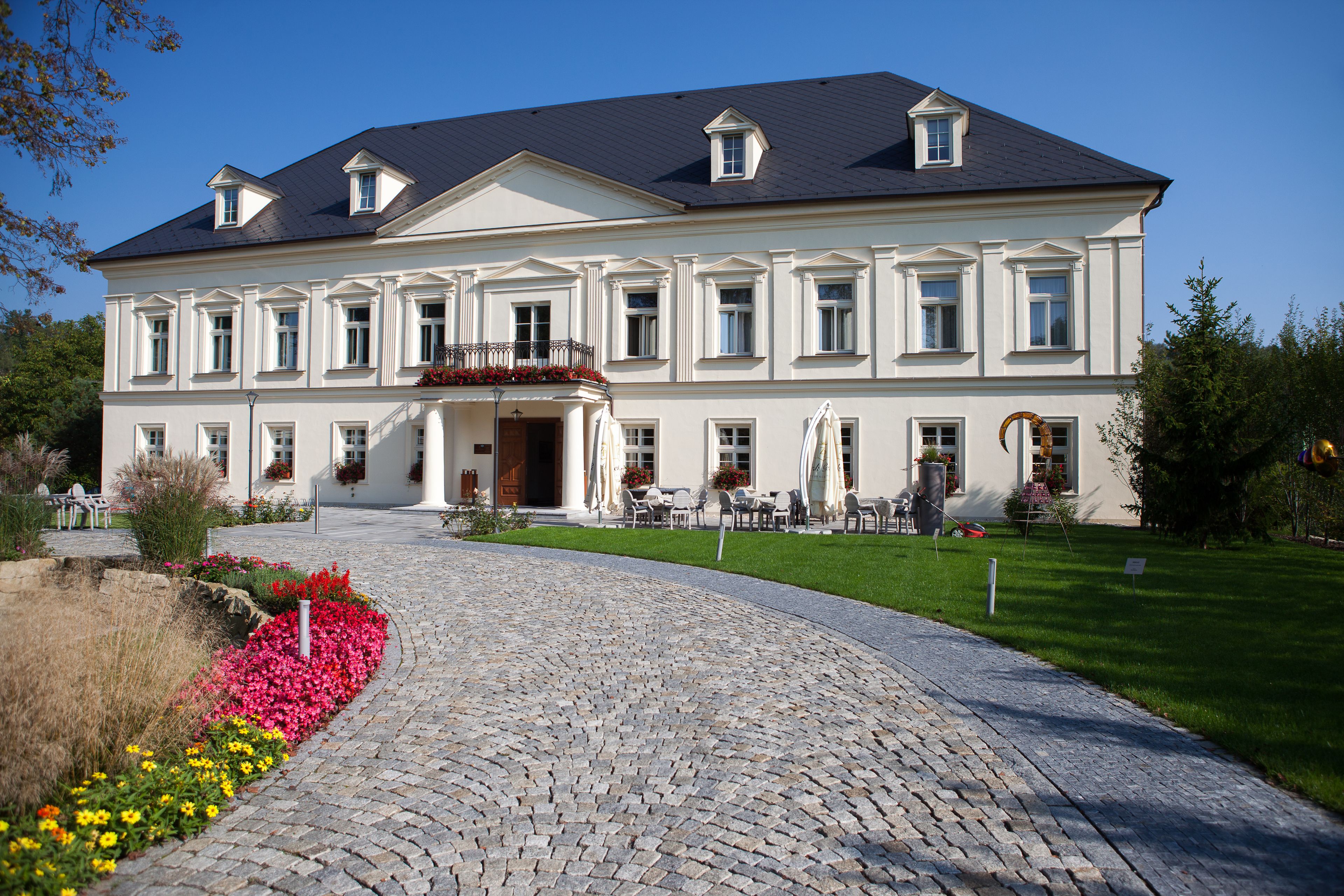A large, two-story building with a symmetrical facade, a central entrance with a balcony, and a paved driveway leading to a seating area with outdoor furniture.