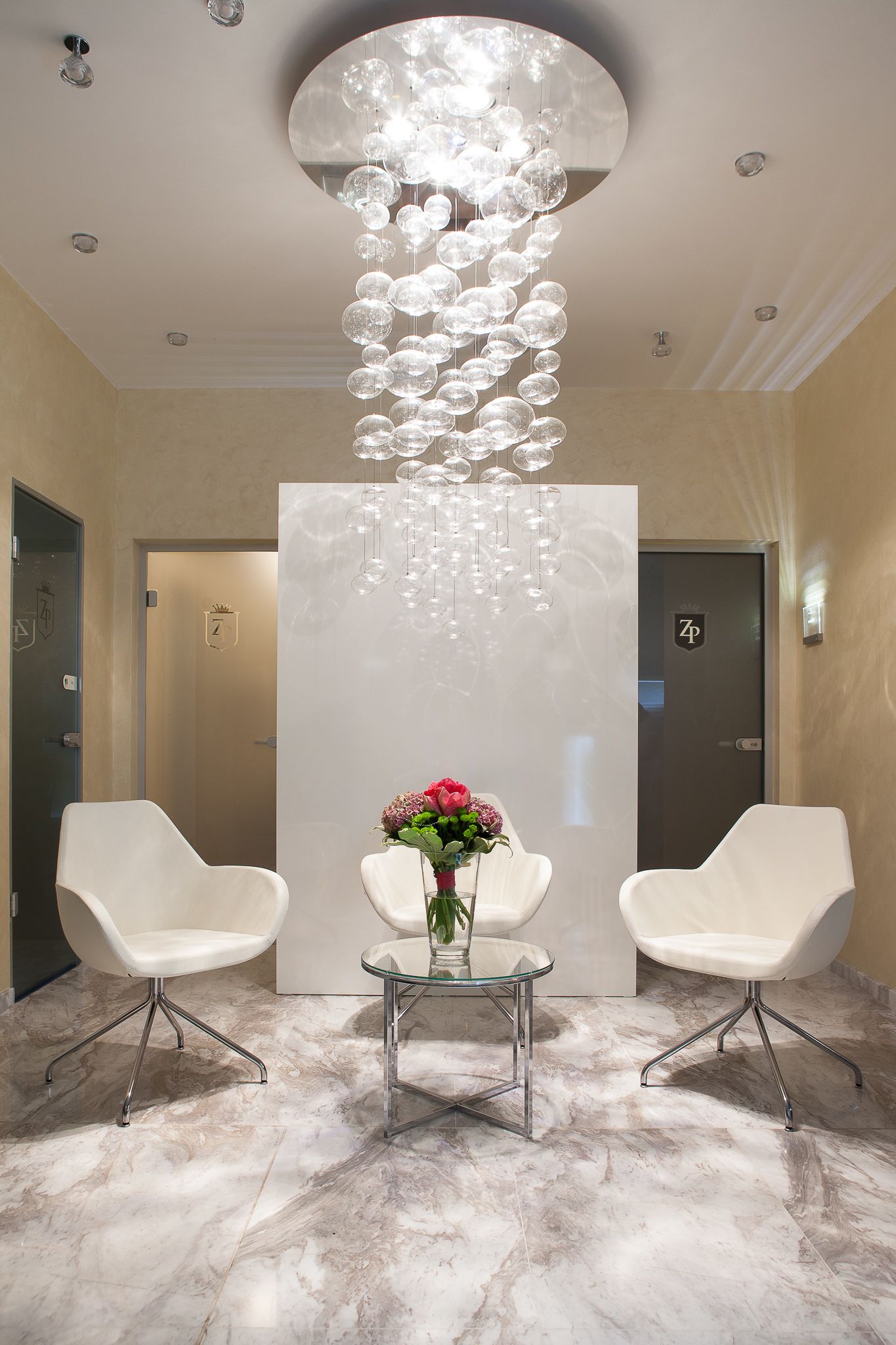 A modern lobby with a marble floor, two white chairs, a glass table with a vase of flowers, and a unique spherical chandelier.
