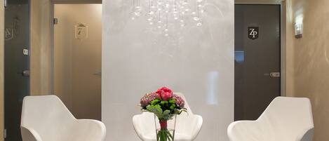 A modern lobby with a marble floor, two white chairs, a glass table with a vase of flowers, and a unique spherical chandelier.