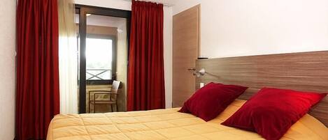 A hotel room with a bed, red curtains, a wooden headboard, and a view through an open door.