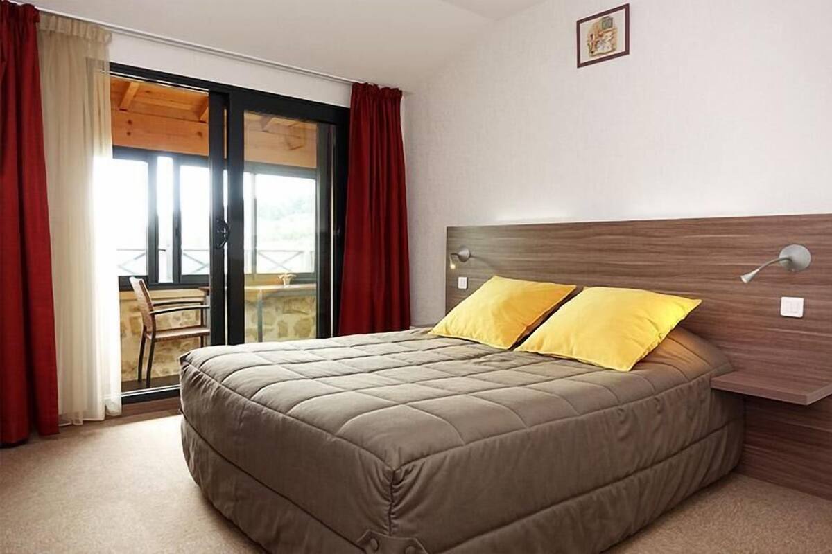 A bedroom with a bed, a wooden headboard, a window with red curtains, and a view of a balcony.