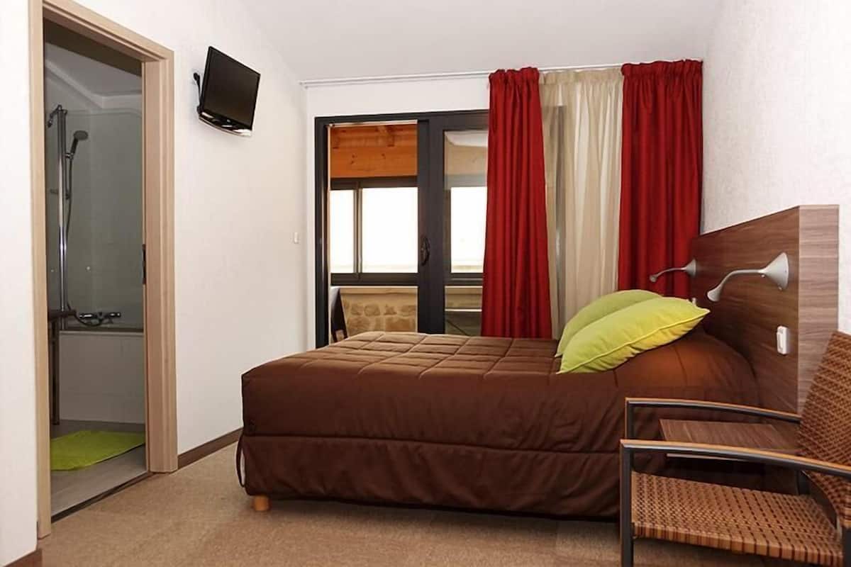A hotel room with a bed, a chair, a TV, a shower, and a window with curtains.
