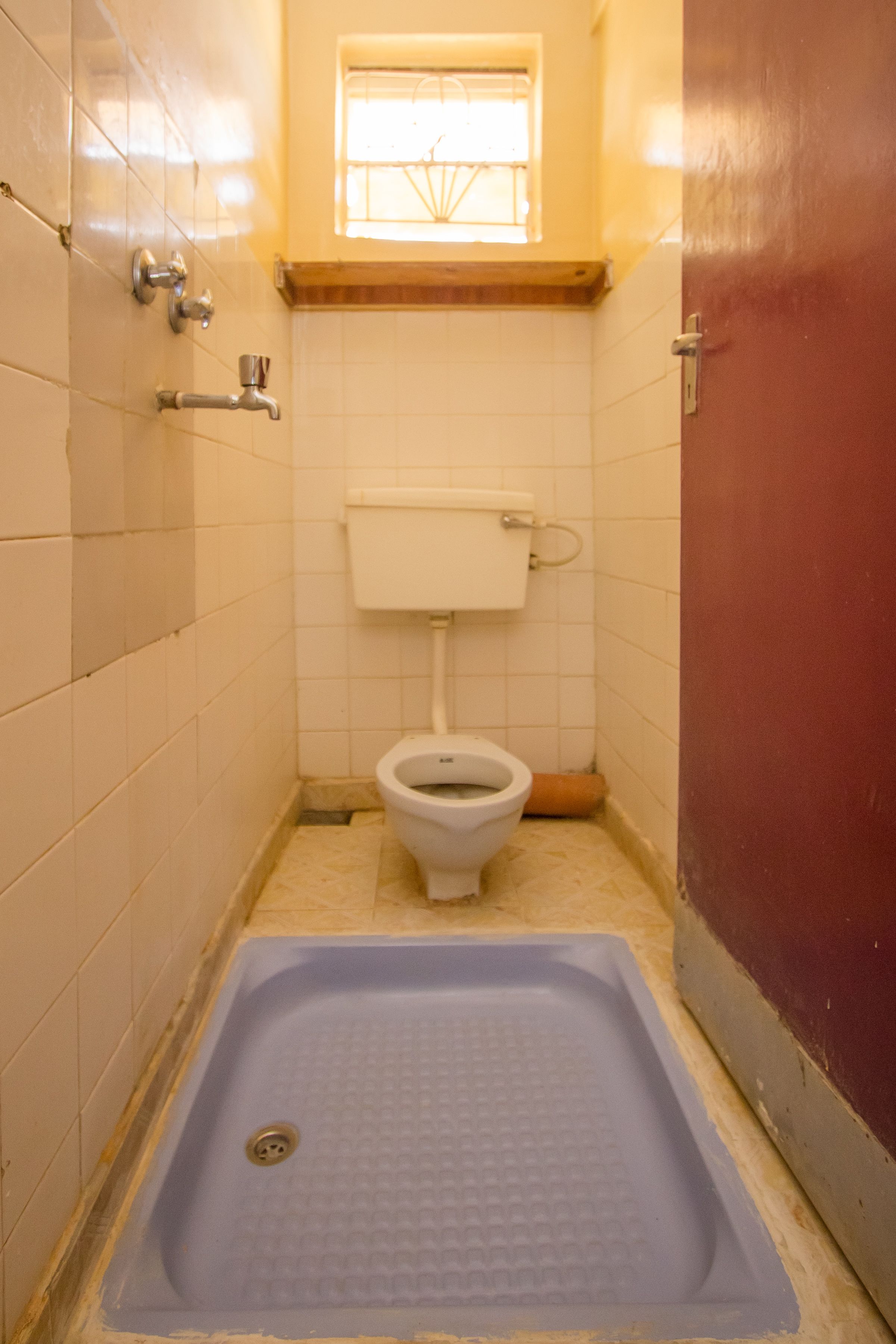 A small, narrow bathroom with a toilet, a blue floor drain, and a window with bars.