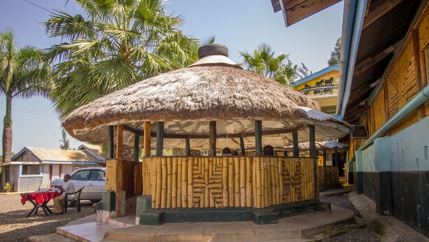 A thatched-roof gazebo with a wooden fence, surrounded by palm trees and a building with a blue and yellow facade. - Jamindas Paradise Motel (Kakamega)