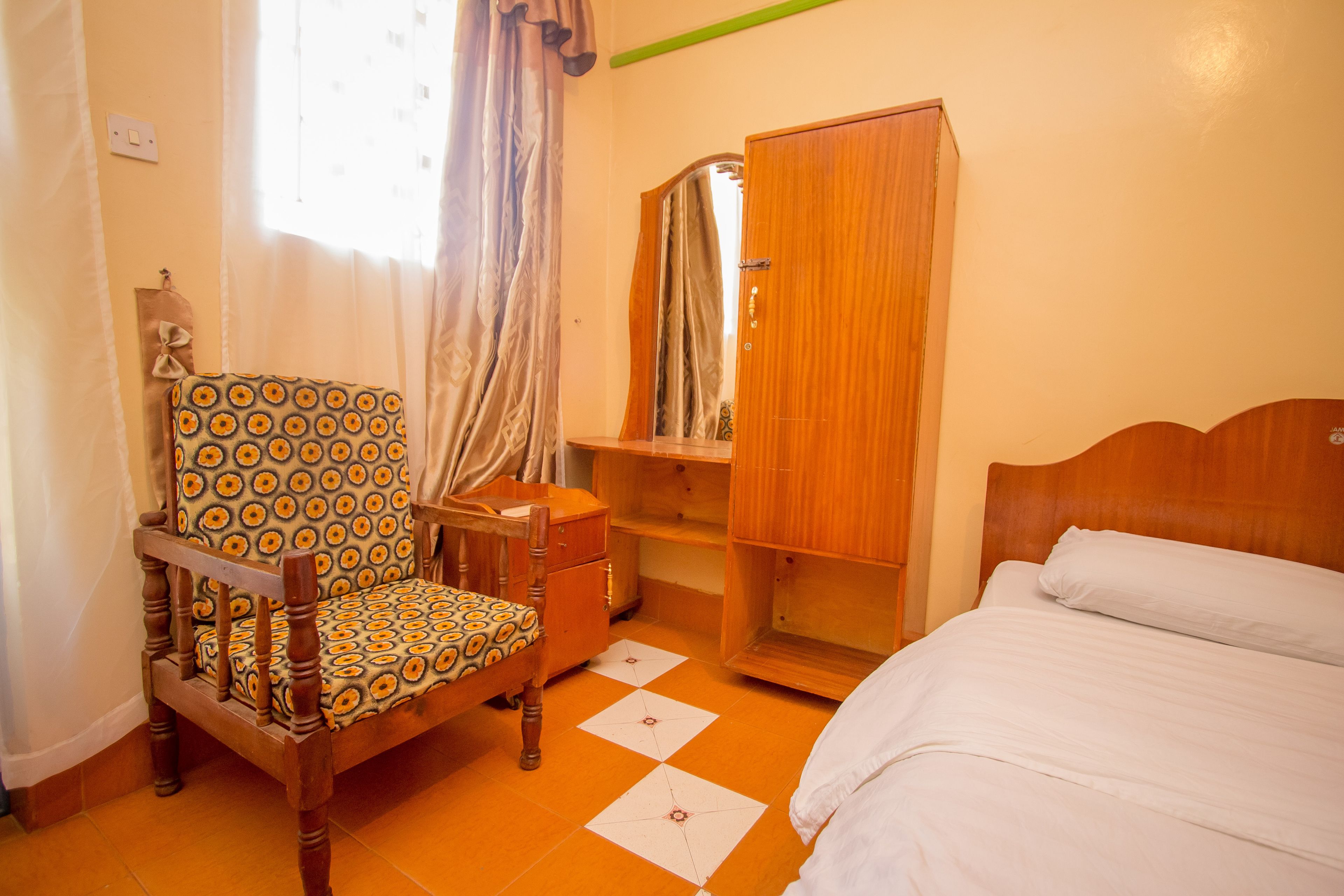 Un dormitorio con una cama de madera, una silla con cojín estampado, un armario de madera y un espejo en la pared.