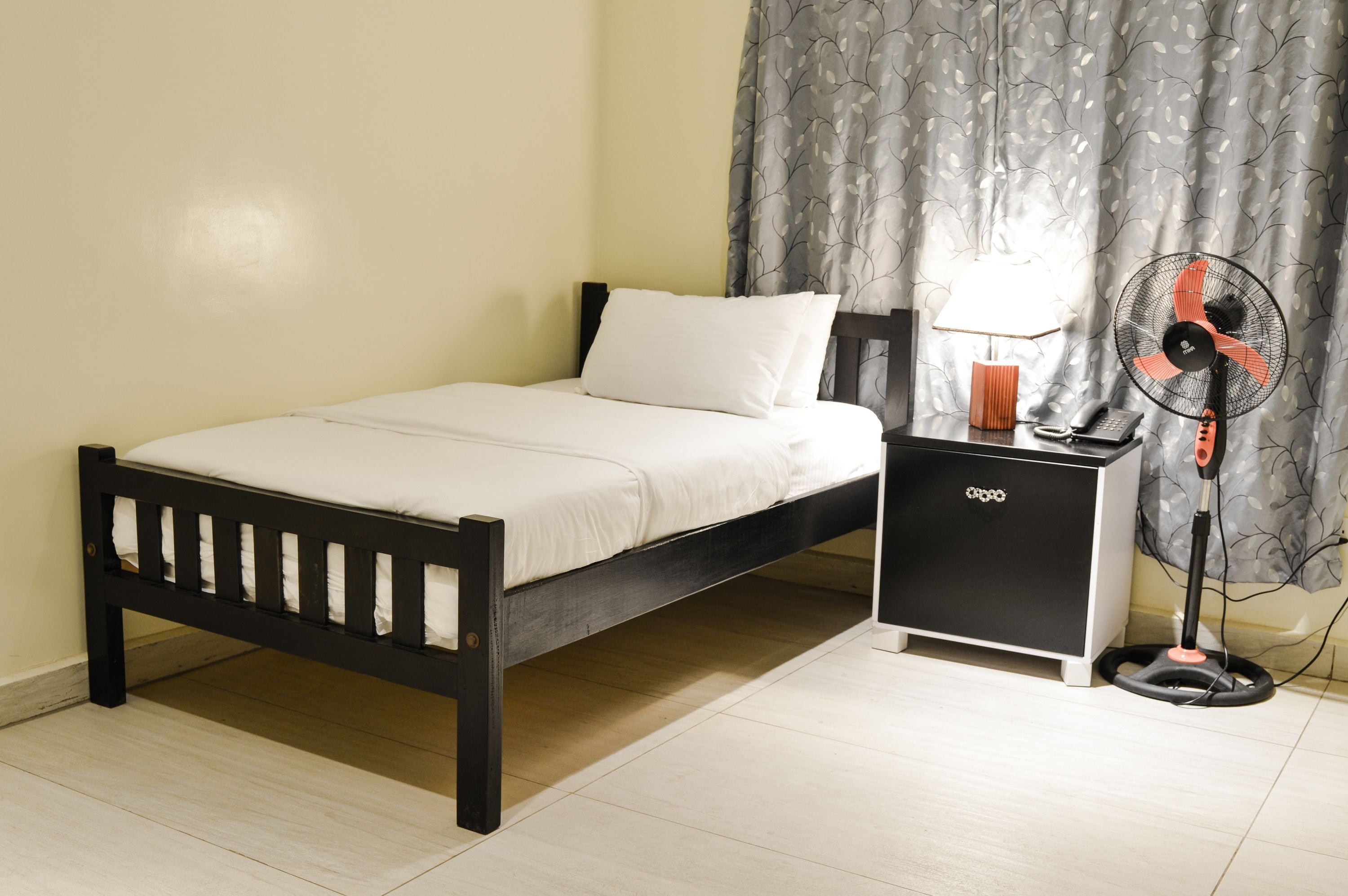Un lit avec un matelas blanc et un cadre noir, une petite table de chevet noire avec une lampe, et un ventilateur sur pied.