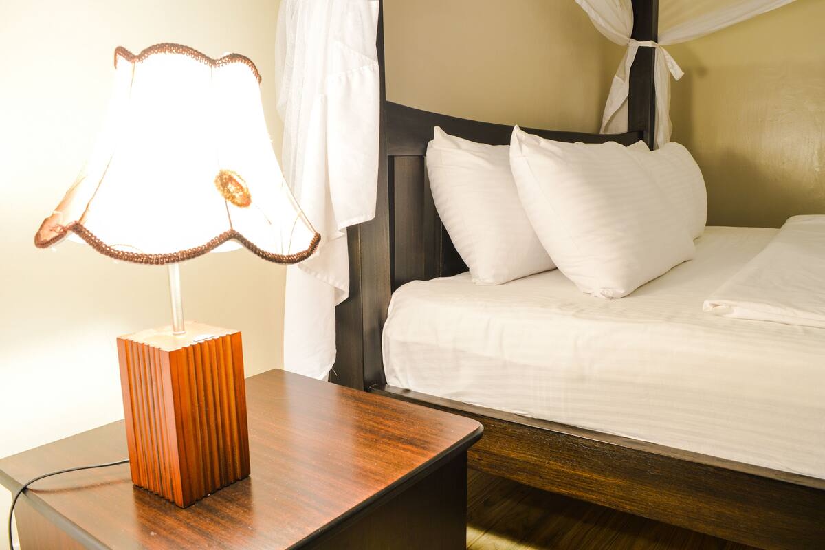 A neatly made bed with white linens and pillows, a wooden bedside table with a lamp, and a dark headboard.