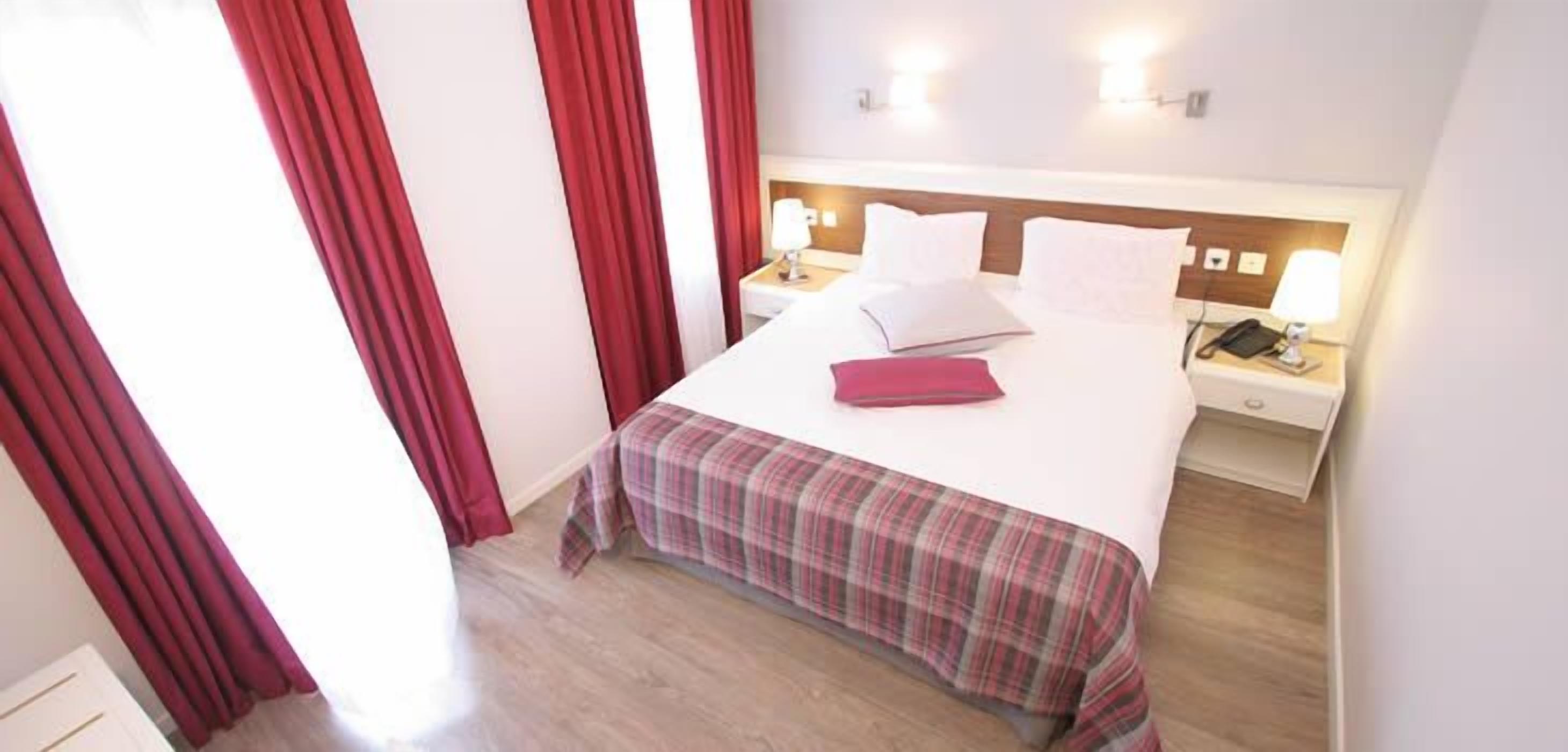 A hotel room with a bed, a nightstand, a lamp, and red curtains.