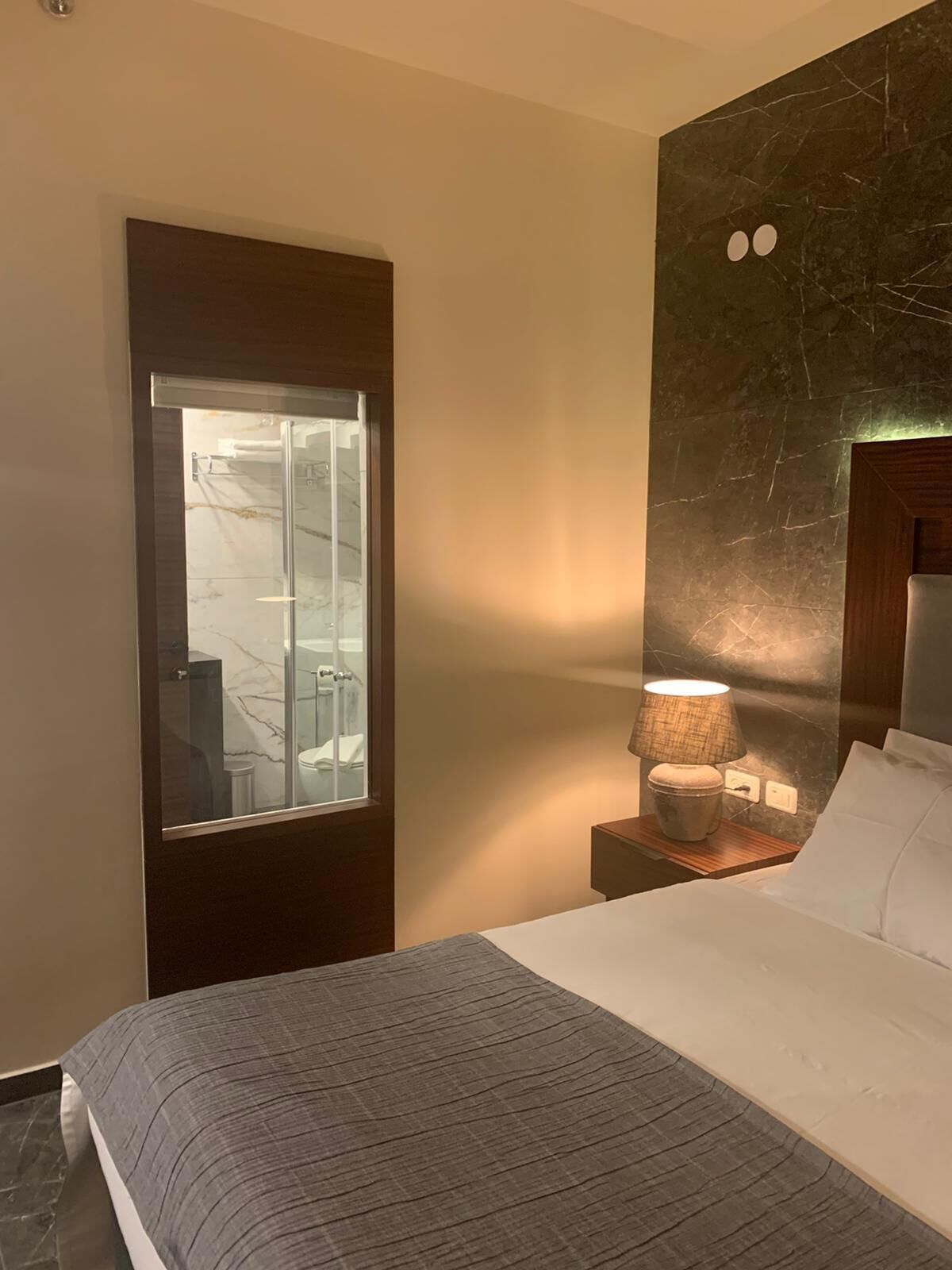 Ein Hotelzimmer mit Bett, Nachttisch mit Lampe, Tür mit Milchglasfenster und Marmorwand mit Holz-Kopfteil.