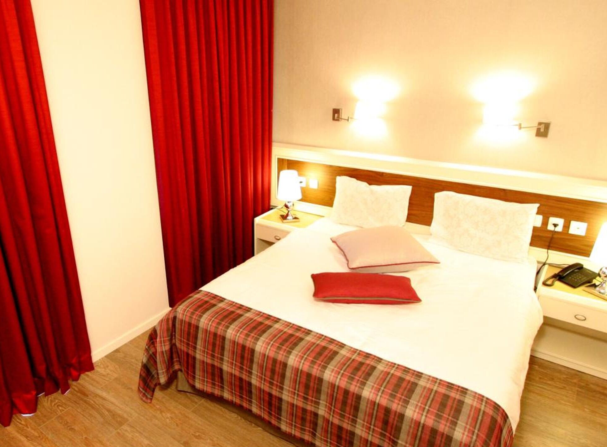 A hotel room with a bed, red curtains, a nightstand with a lamp, and a phone.