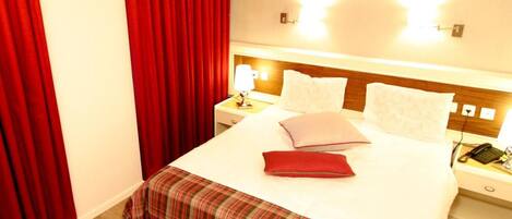 A hotel room with a bed, red curtains, a nightstand with a lamp, and a phone.