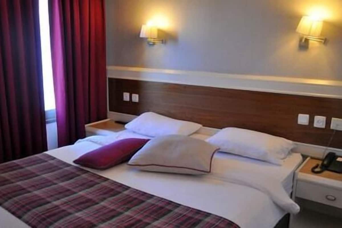 Une chambre d’hôtel avec un lit, deux tables de chevet, une petite table, une lampe et une fenêtre avec des rideaux.