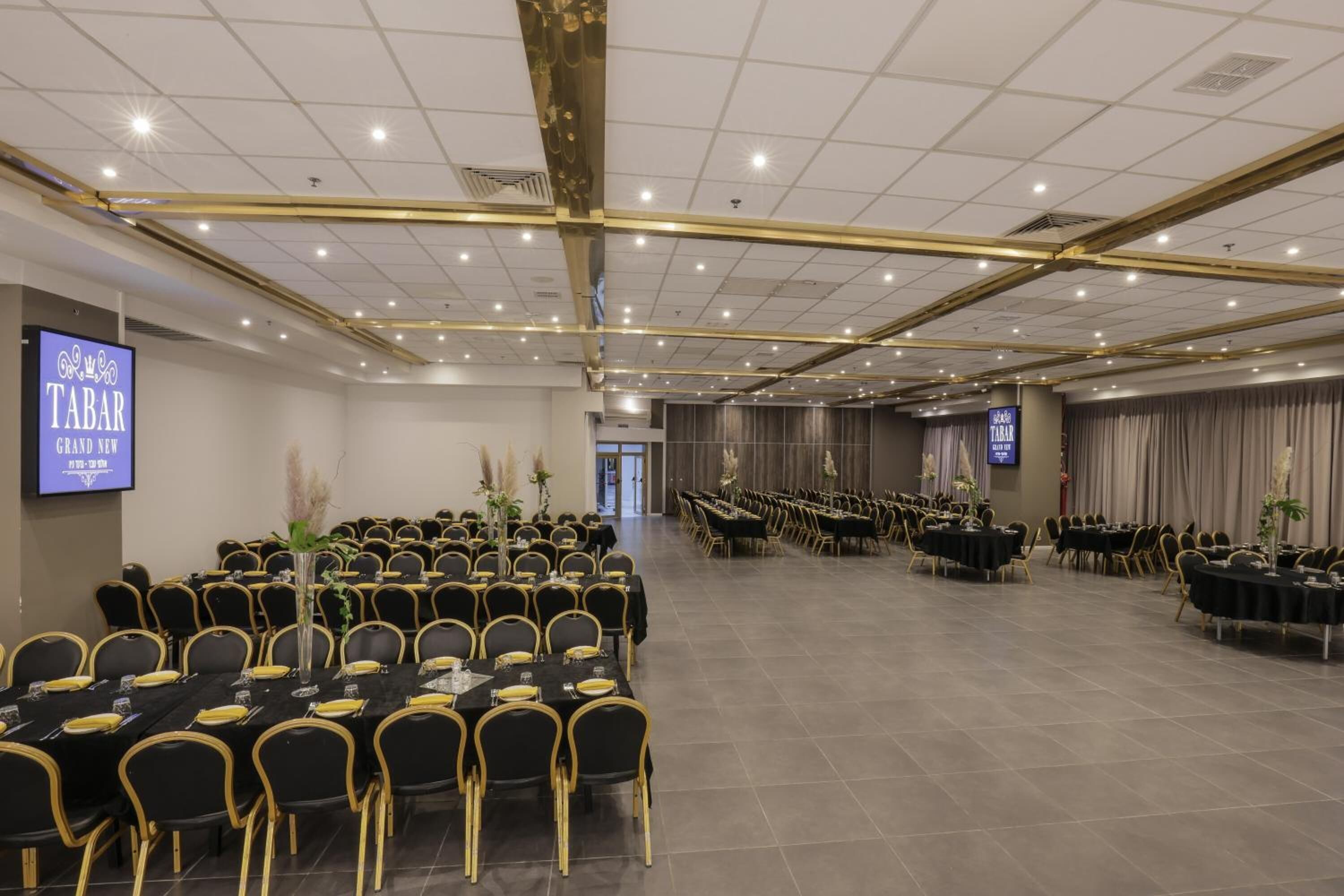Une grande salle de conférence avec de nombreuses rangées de chaises et de tables noires, un écran de projection et une enseigne indiquant « TABAR GRAND NEW ».