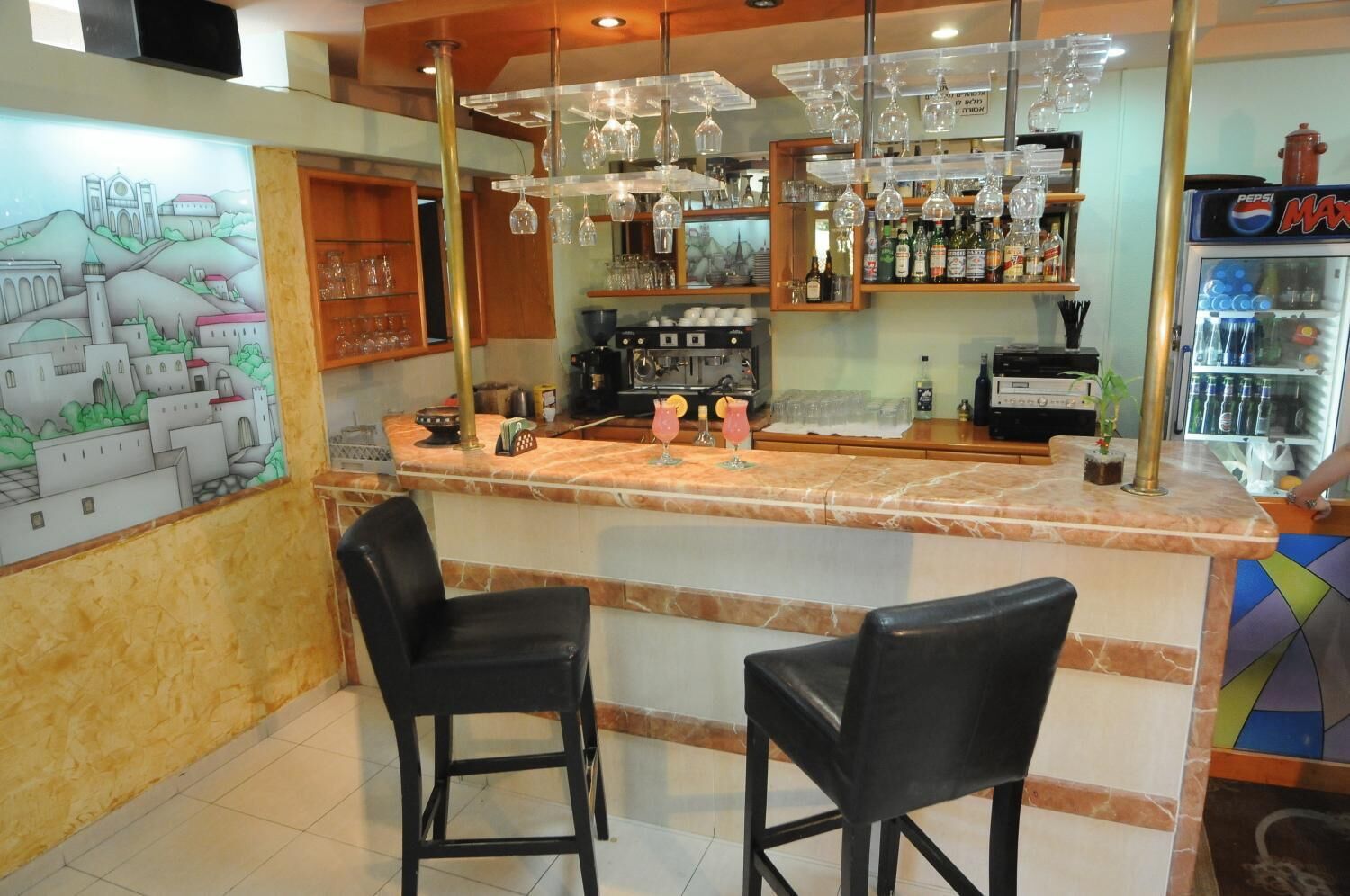A bar area with two black bar stools, a counter with a marble top, shelves with bottles and glasses, and a mural on the wall.