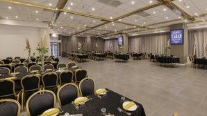 Une vaste salle de banquet, avec de longues rangées de tables et de chaises, une scène et un grand écran sur lequel est affiché le message « TABAR GRAND NEW ».