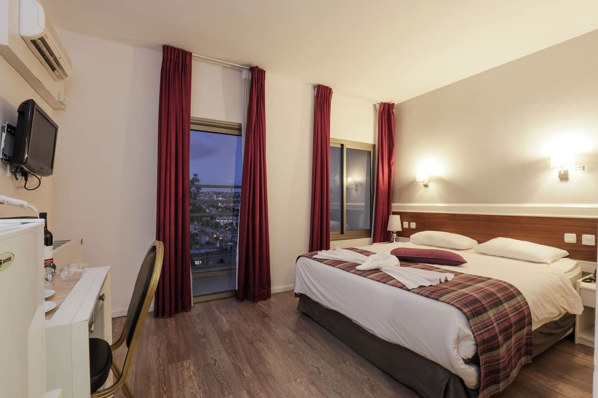 Une chambre d’hôtel comprenant un lit, un bureau, une chaise, un appareil de climatisation, une télévision et une fenêtre avec des rideaux.