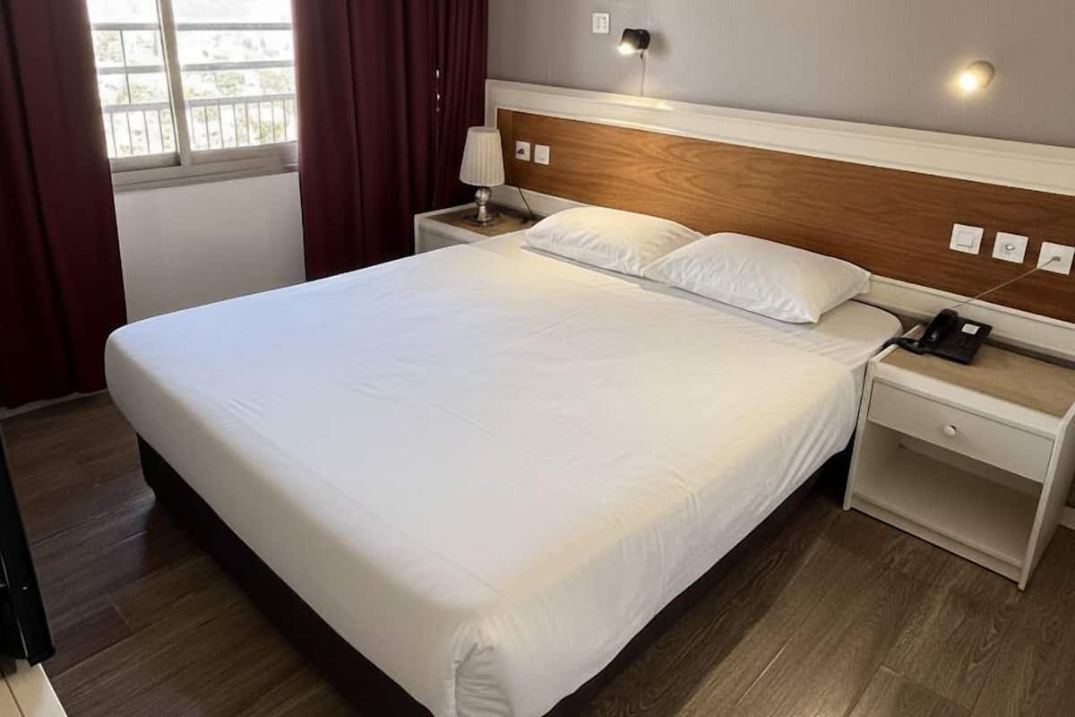 Une chambre d’hôtel avec un grand lit, une tête de lit en bois, une table de chevet avec une lampe et une fenêtre avec des rideaux.