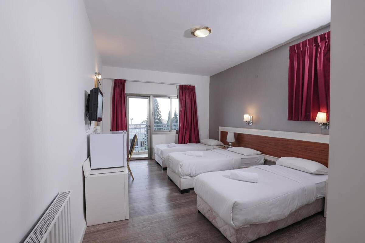 Une chambre d’hôtel avec trois lits, un bureau, une télévision et une fenêtre avec des rideaux.