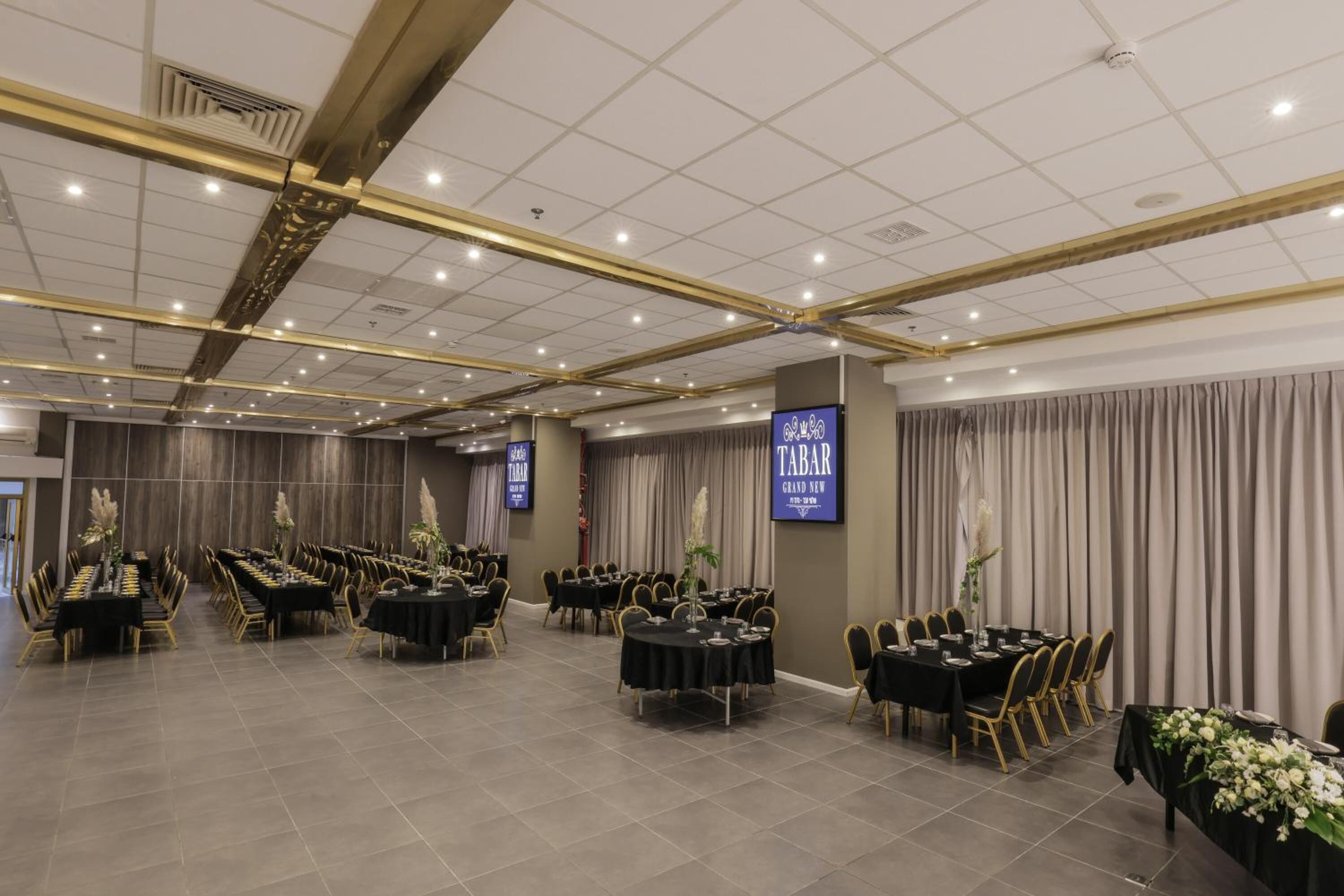 A banquet hall with multiple round tables and chairs, a stage area, and a large screen displaying the name "TABAR".