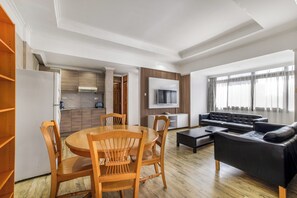 A modern living room with a dining area, a black leather sofa, wooden chairs, a TV mounted on the wall, and large windows. - Kates Apartments (Nairobi)