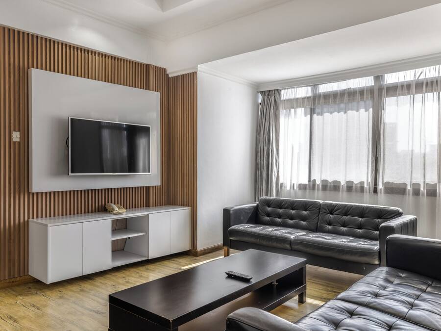 A modern living room with a flat-screen TV mounted on a white wall, a leather sofa, a coffee table, and a wooden accent wall.