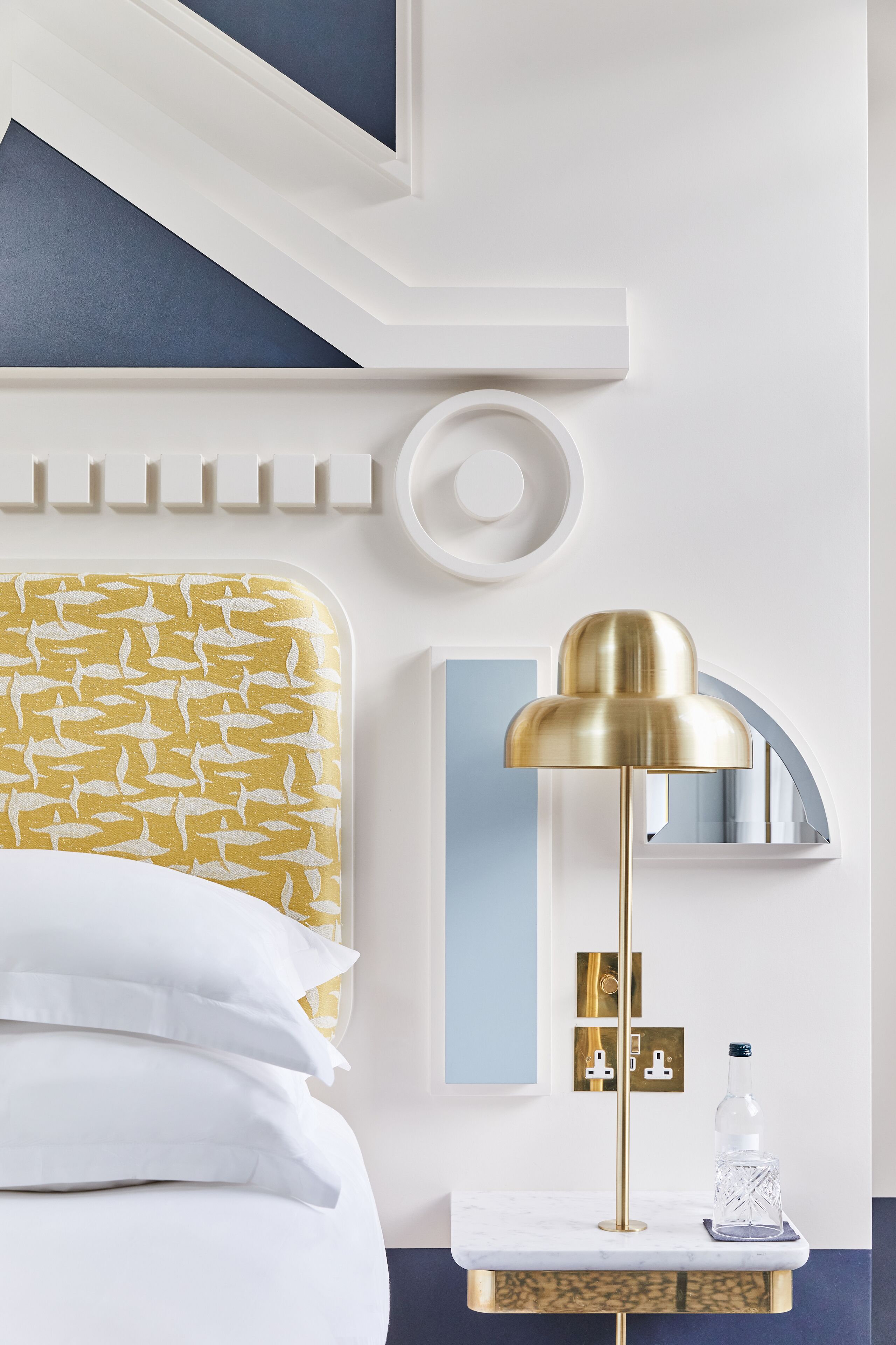 A hotel room with a bed, a bedside table with a lamp, a mirror, and a wall-mounted control panel.
