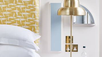 A hotel room with a bed, a bedside table with a lamp, a mirror, and a wall-mounted control panel.