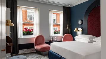 Quarto de hotel com cama, duas cadeiras, uma mesinha com uma taça e garrafa de vinho, e vista para um edifício de tijolos vermelhos através de duas janelas.