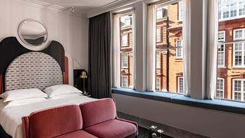 Habitación de hotel con cama, sofá rojo, espejo redondo y vista a un edificio de ladrillo a través de la ventana.