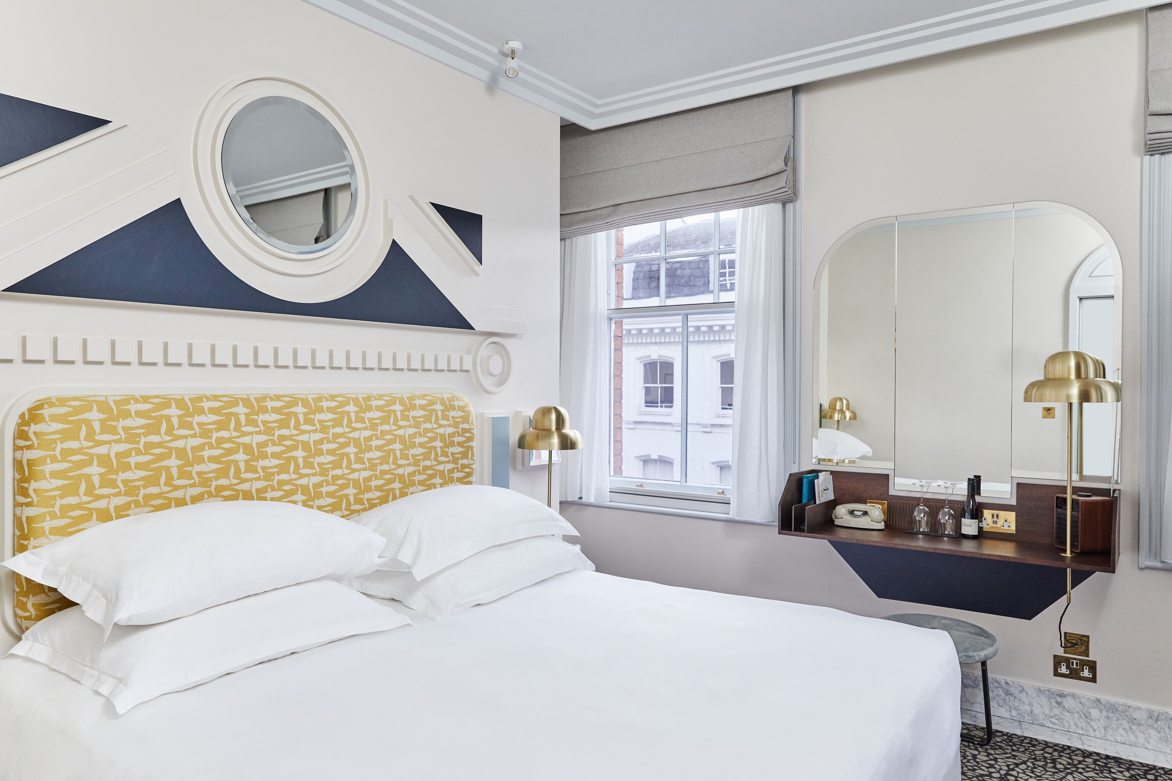 A hotel room with a bed, a round mirror, a dresser with a mirror, and a window with a view of buildings.