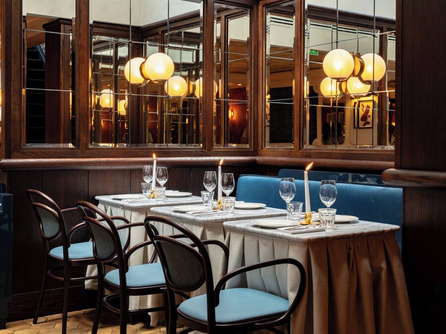 Un restaurante con mesas dispuestas para comer, sillas y un amplio ventanal con iluminación decorativa.