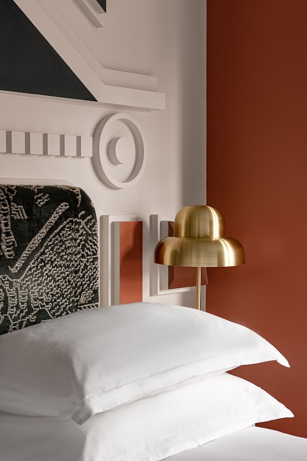 Un dormitorio con una cama, almohadas blancas, un plaid estampado, una lámpara dorada y una pared con diseño geométrico.