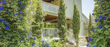 A two-story house with a balcony, surrounded by lush greenery and blue flowers.