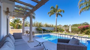 Zona junto a la piscina con un espacio de descanso techado, piscina y abundante vegetación.