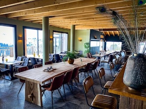 A restaurant with wooden tables and chairs, large windows, and a decorative plant.