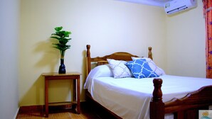A bedroom with a wooden bed, white bedding, a small wooden nightstand, a potted plant, and an air conditioning unit.