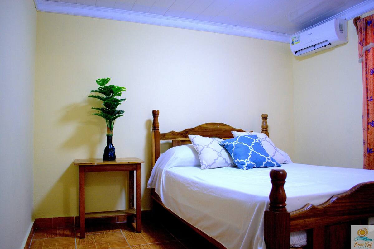 A bedroom with a wooden bed, white bedding, a small wooden nightstand, a potted plant, and an air conditioning unit.