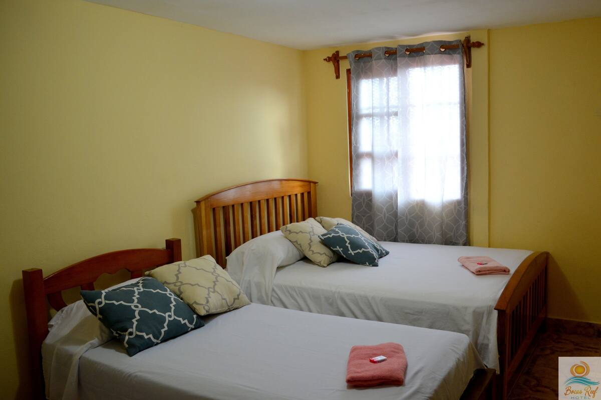 A room with two single beds, each with a wooden headboard, white bedding, and patterned pillows. A window with a curtain is visible, and the walls are painted yellow.