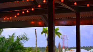 A view from a covered outdoor area with string lights, palm trees, and buildings in the background.