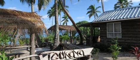 A rustic wooden cabin with a thatched roof and a sign reading "Teukona".