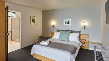 A neatly made bed with a grey and white duvet, a green pillow, and a folded towel on the side. There are two wall-mounted lights and a small bedside table with a phone and charger.