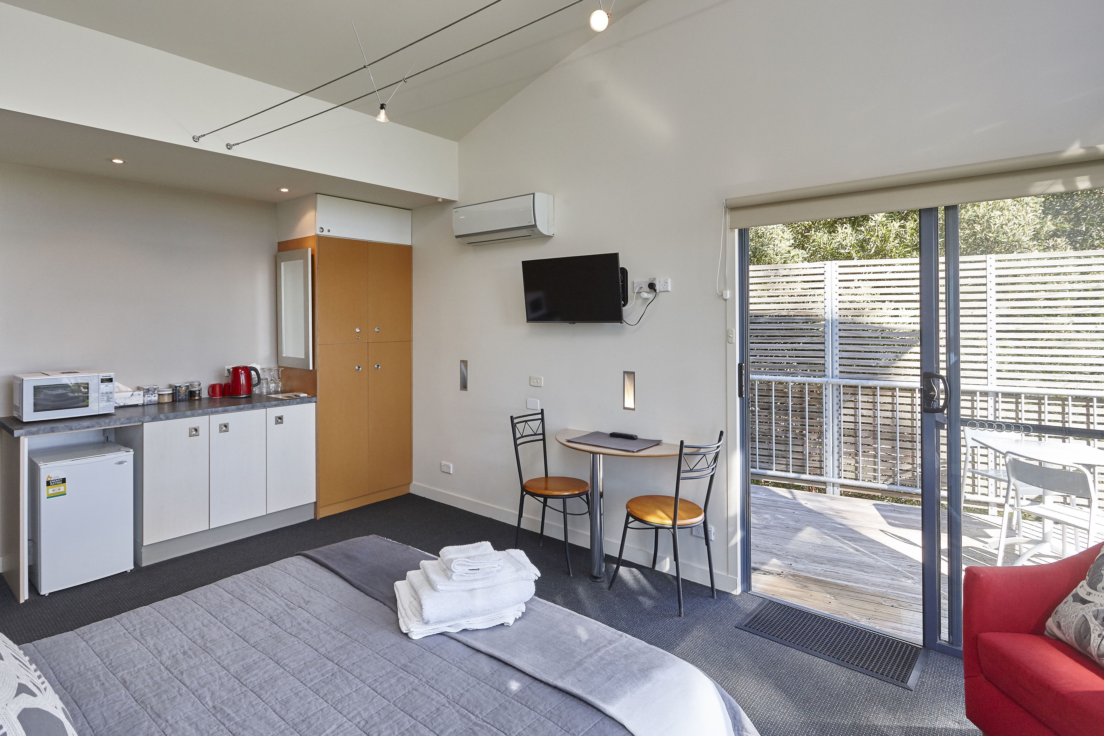 A hotel room with a bed, a desk with a chair, a small kitchenette, and a TV mounted on the wall.