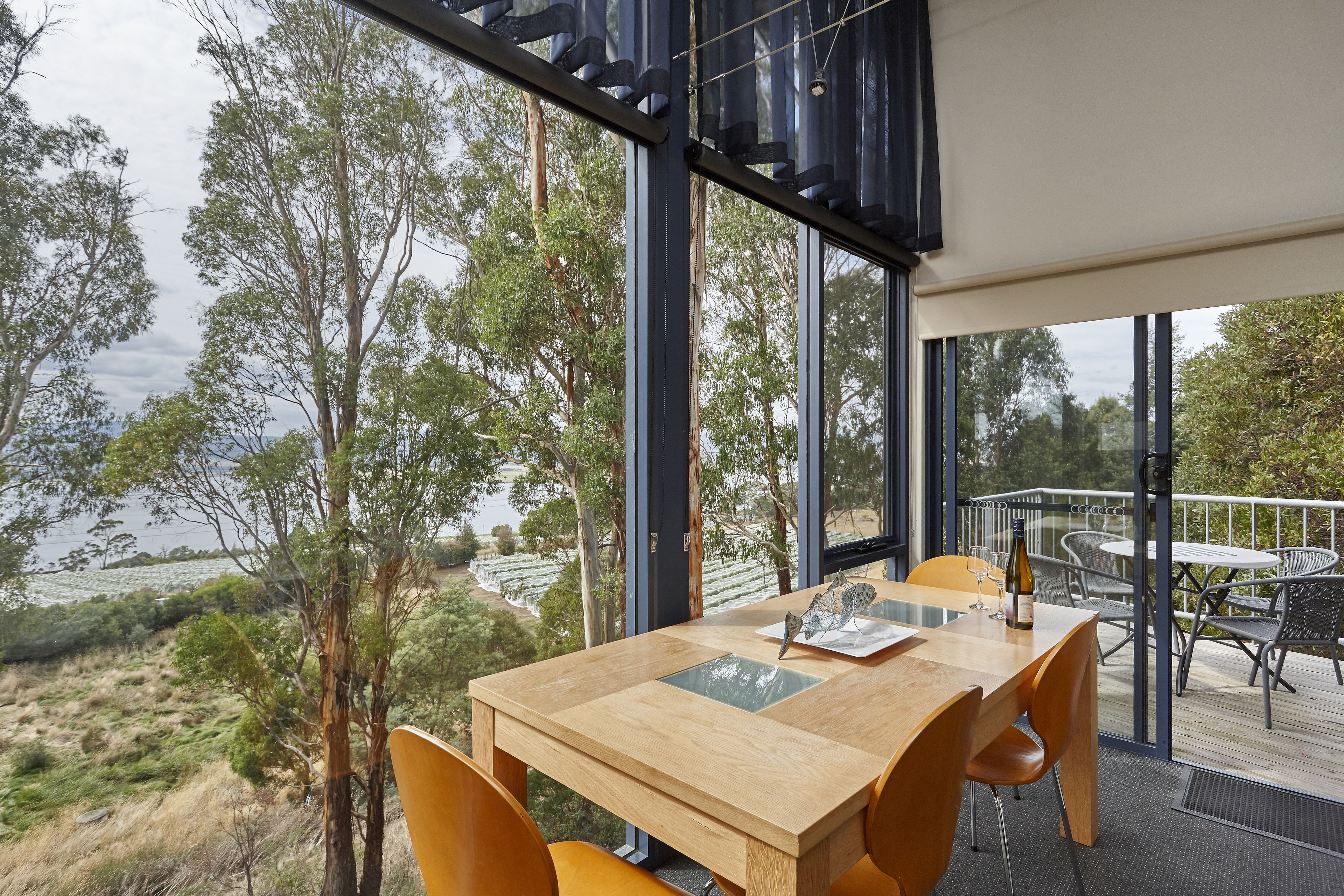 Un comedor con mesa y sillas de madera, ventanas amplias que ofrecen vistas a árboles y un cuerpo de agua.