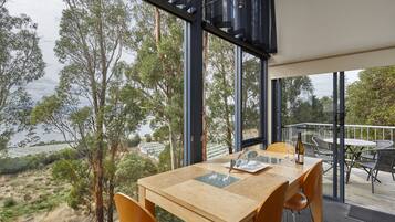 Un comedor con mesa y sillas de madera, ventanas amplias que ofrecen vistas a árboles y un cuerpo de agua.