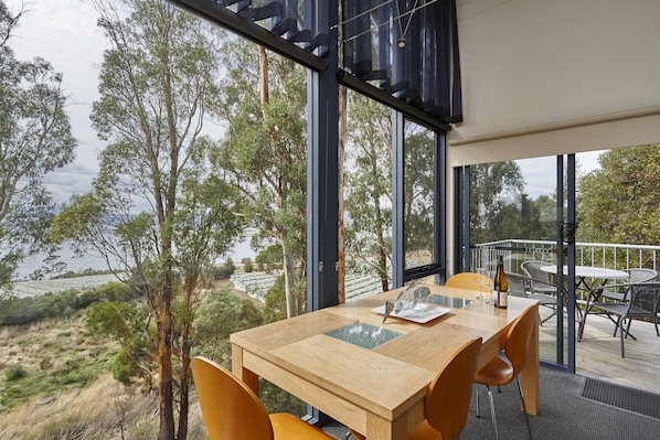 Un comedor con mesa y sillas de madera, ventanas amplias que ofrecen vistas a árboles y un cuerpo de agua.