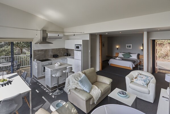 Un área de estar y comedor moderna, de planta abierta, con kitchenette, un dormitorio con cama y mesita de noche, y un balcón con barandilla.