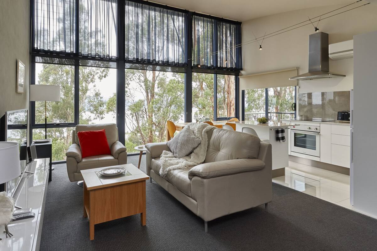 A modern living room with a sofa, coffee table, and a dining area with a table and chairs. A kitchen with appliances is visible in the background.