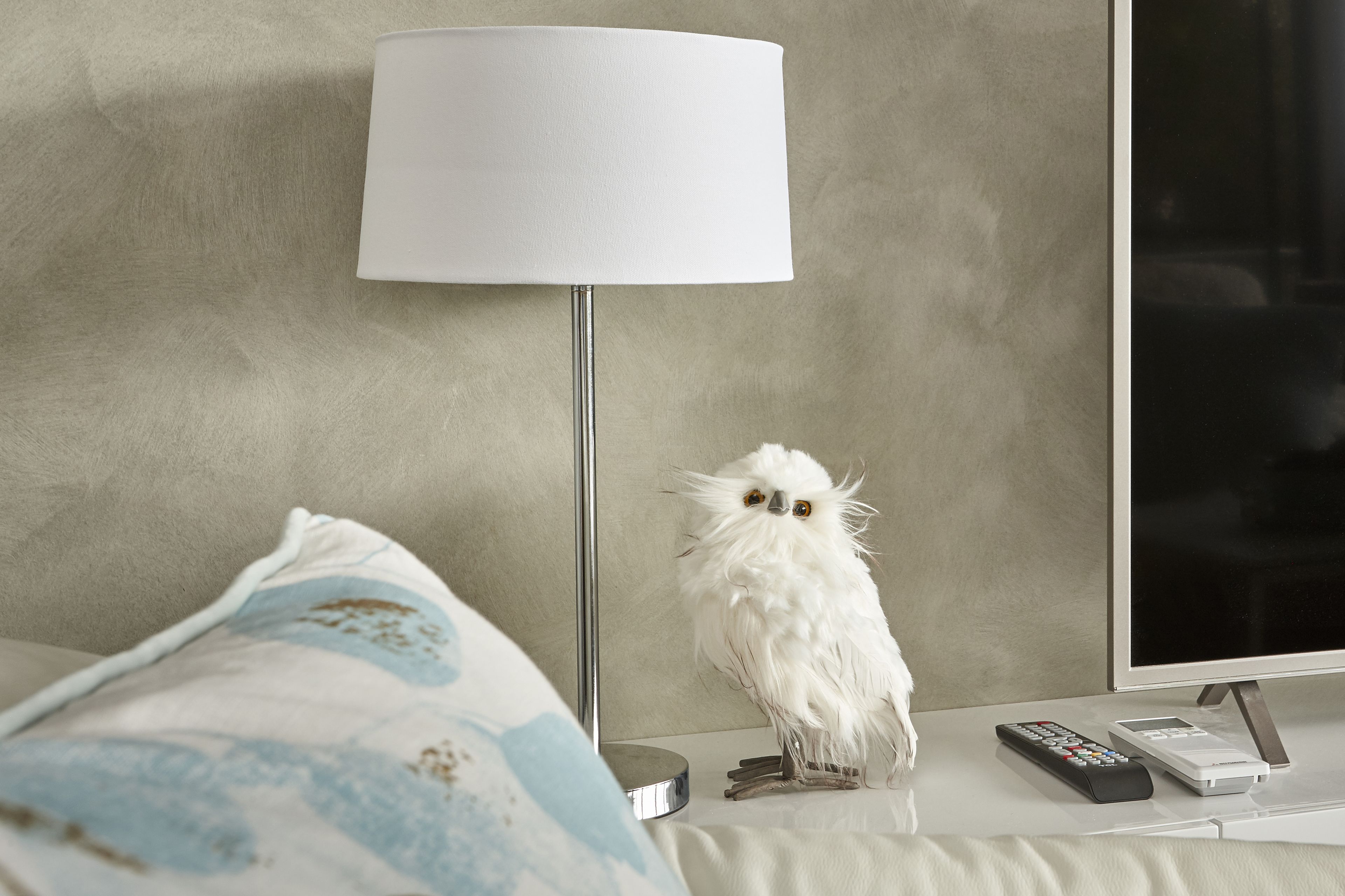 A white fluffy owl on a bedside table next to a lamp and remote controls.